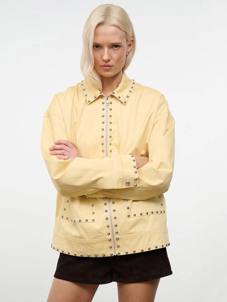 A person with blonde hair wears the STAUD Clarke Jacket in Butter Studs and dark shorts, standing against a plain white background with arms crossed.