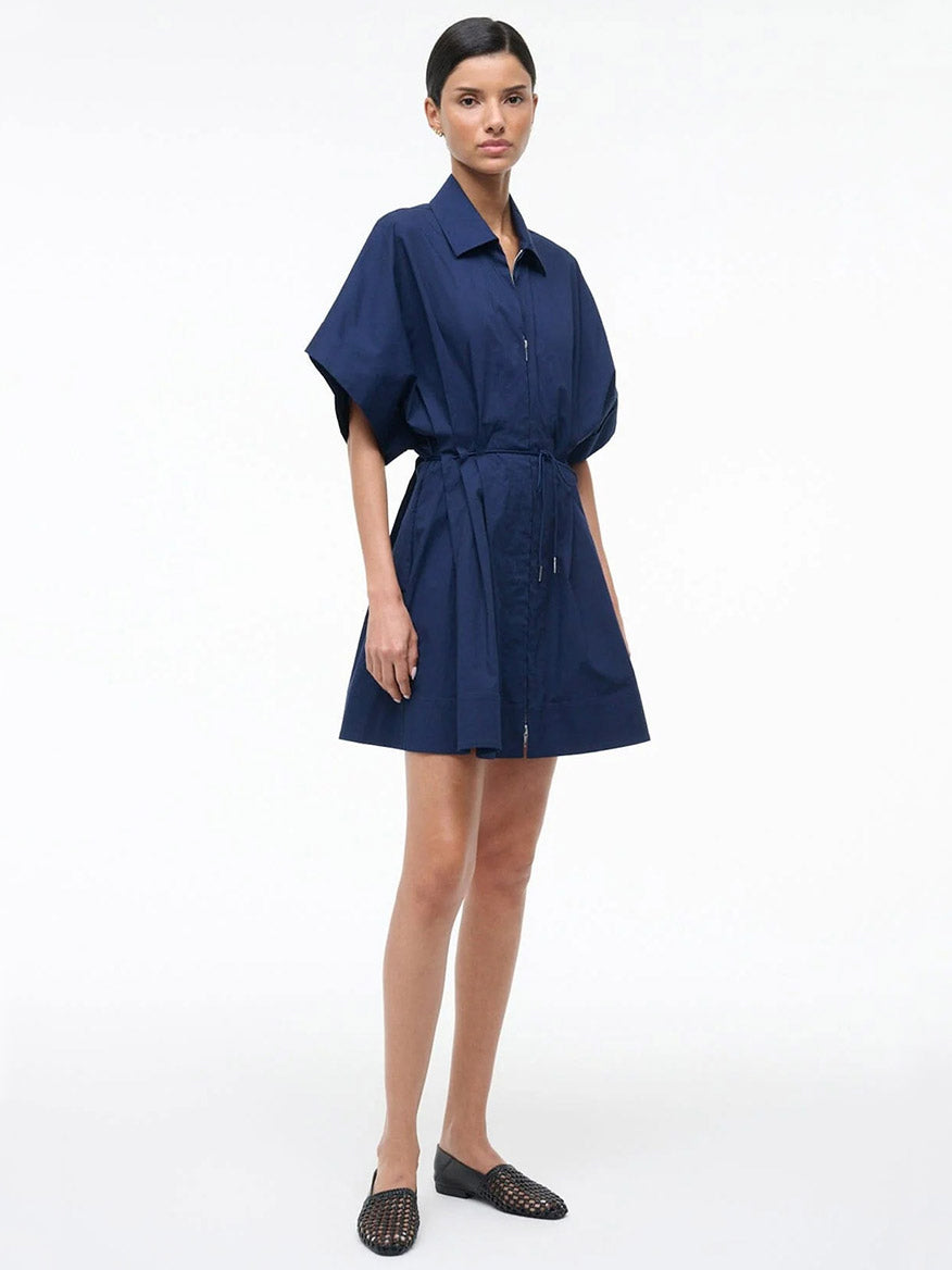 A woman stands against a white background wearing the STAUD Cori Dress in Navy, a collared, short-sleeve dress with an adjustable drawstring waist, paired with black woven flats.