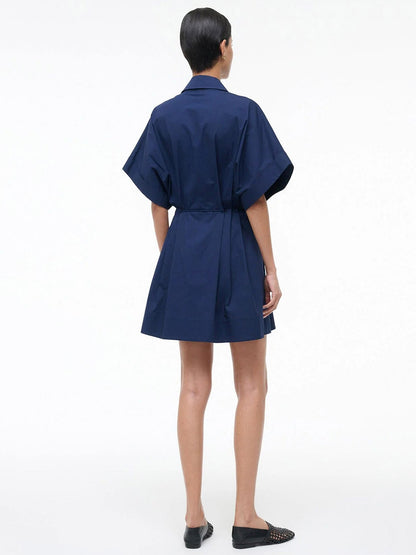 A person stands with their back to the camera in a STAUD Cori Dress in Navy—short-sleeved, collared, with an adjustable drawstring waist—and black flat shoes, set against a plain white background.