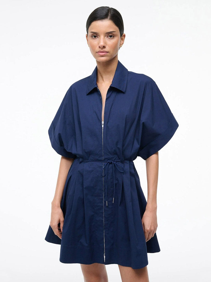 A woman poses against a plain background wearing the STAUD Cori Dress in Navy, which features short sleeves, a front zipper, a collared neckline, and an adjustable drawstring waist.