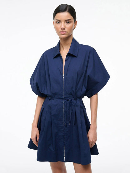 A woman poses against a plain background wearing the STAUD Cori Dress in Navy, which features short sleeves, a front zipper, a collared neckline, and an adjustable drawstring waist.