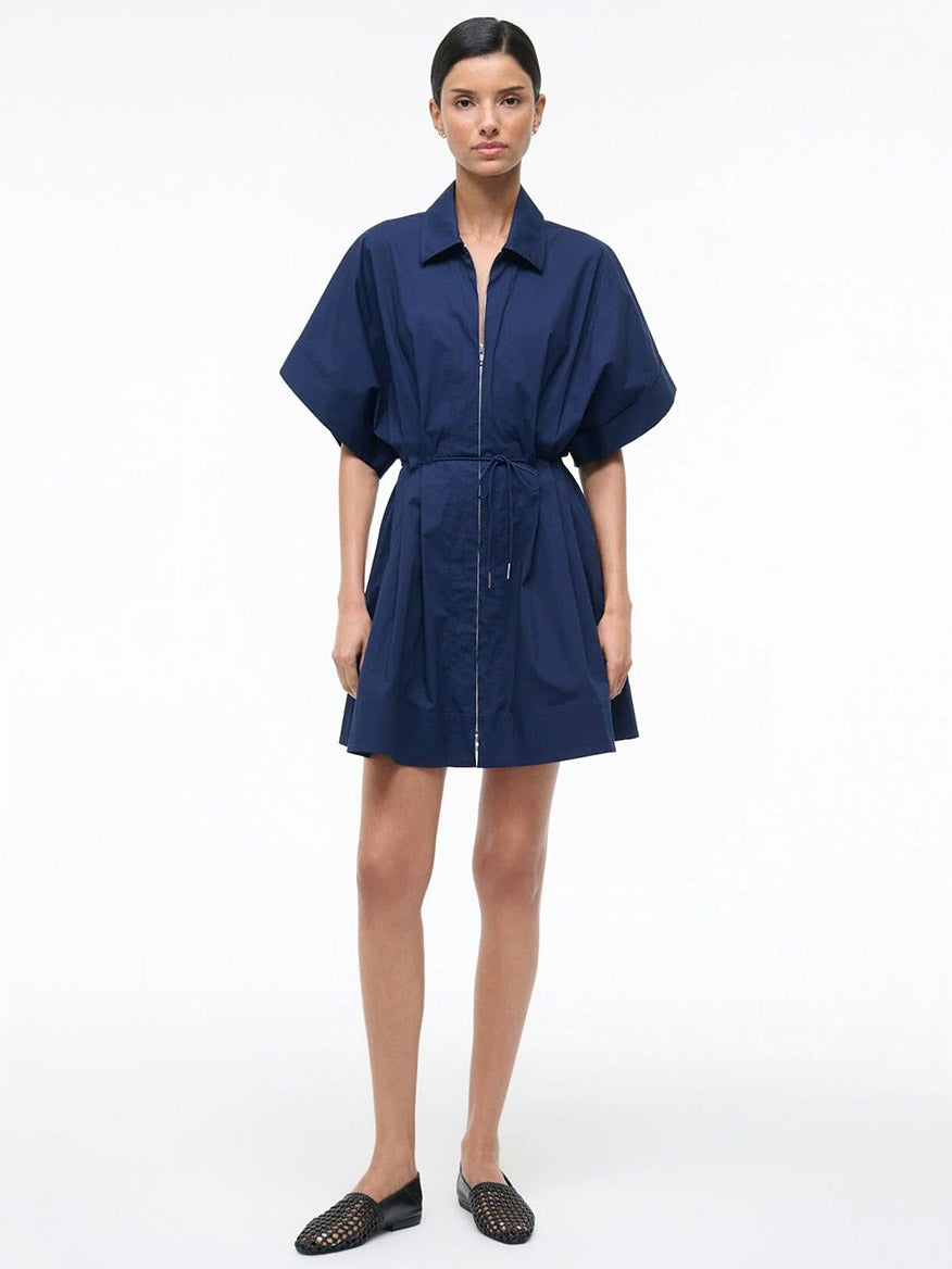 A woman stands against a plain background wearing the STAUD Cori Dress in Navy, which features a collared neckline and an adjustable drawstring waist, paired with black woven flats.