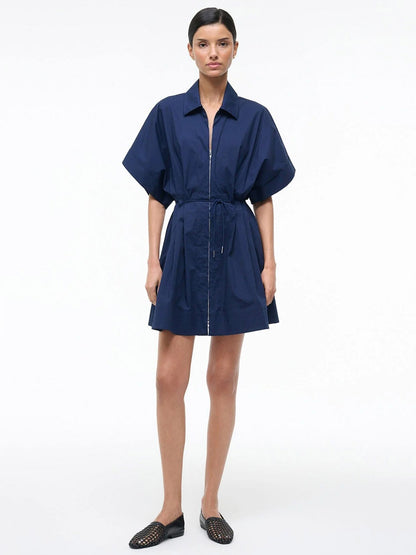 A woman stands against a plain background wearing the STAUD Cori Dress in Navy, which features a collared neckline and an adjustable drawstring waist, paired with black woven flats.