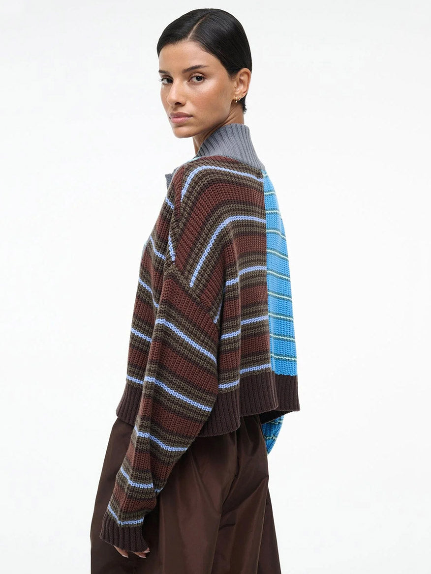 A person wearing the STAUD Cropped Hampton Sweater in Zuma Multi, a chunky striped knit with long sleeves, and loose brown pants, stands against a plain white background.