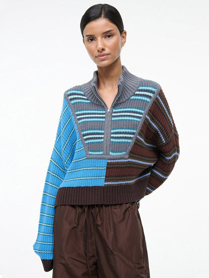 A woman stands against a plain background, wearing the STAUD Cropped Hampton Sweater in Zuma Multi, featuring a chunky cotton knit patchwork design in blue and brown tones, paired with loose brown pants.