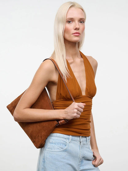A woman with straight blonde hair wears a sleeveless brown top with an asymmetrical silhouette and light blue jeans, carrying the STAUD Dani Shoulder Bag in Tabac, which features a knotted strap, on her shoulder against a plain white background.
