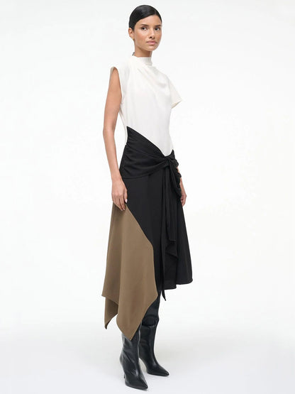 A woman wears the STAUD Davina Dress in Black Multi—a sleeveless, high-neck, asymmetrical dress with a handkerchief hem—styled with black knee-high boots, posed against a plain background.