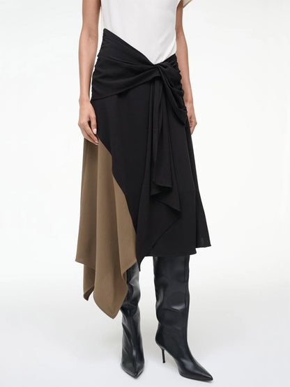 A woman wears the STAUD Davina Dress in Black Multi, featuring a black and brown asymmetrical handkerchief hem and knot detail, paired with black knee-high boots.