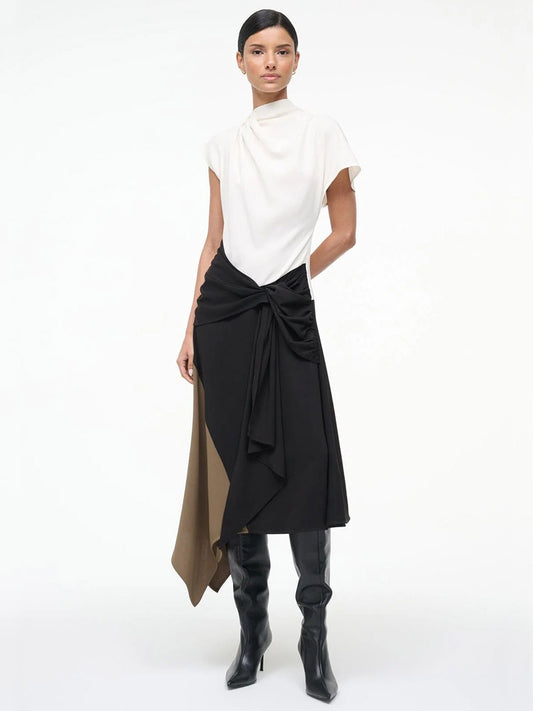 A woman stands against a plain background wearing the STAUD Davina Dress in Black Multi, featuring a high-neck top and color-blocked handkerchief hem, paired with black knee-high boots.
