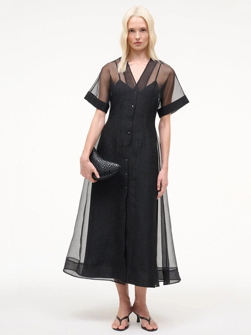 A woman faces forward in the STAUD Elle Dress in Black, layered over a slip, holding a textured black clutch and wearing black sandals, set against a plain white background.