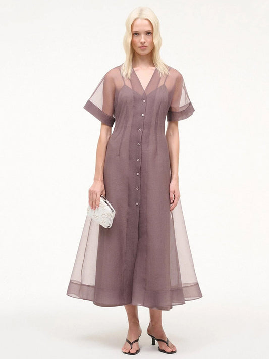A woman in a STAUD Elle Dress in Mink—a sheer short-sleeve A-line dress—holds a white clutch, standing in sandals against a plain background.