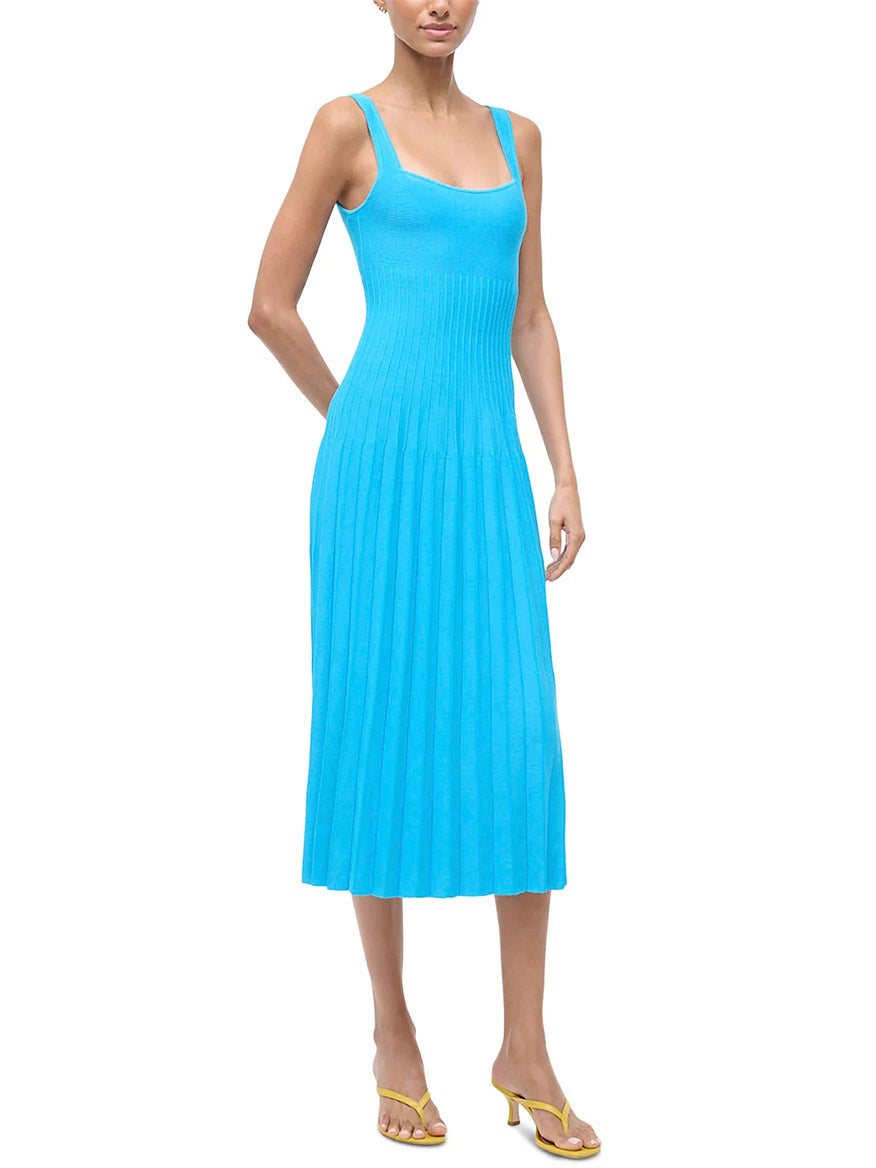 A woman wearing the STAUD Ellison Sleeveless Dress in Pool—a square-neck, bright blue compact knit dress with a pleated skirt—paired with yellow heeled sandals.