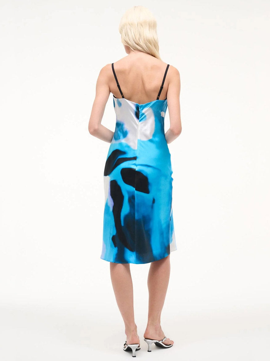 A person with long blond hair, wearing the STAUD Evangeline Silk Dress in Blue Haze and heeled sandals, stands with their back to the camera against a plain white background.