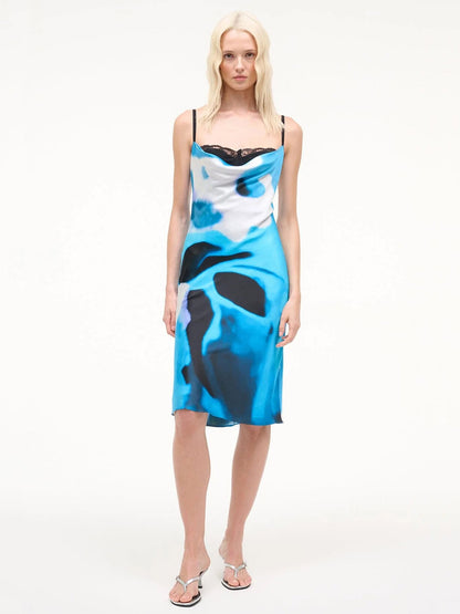 A woman stands facing forward wearing the STAUD Evangeline Silk Dress in Blue Haze, featuring an abstract black and white print, silver sandals, and straight blonde hair.
