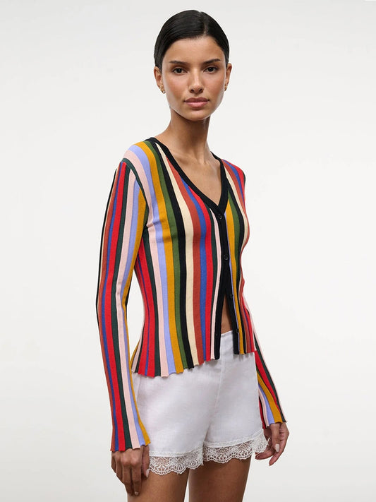 A woman stands against a plain background wearing the STAUD Ezra Cardigan in Artist Stripe with long sleeves, paired with white lace-trim shorts.