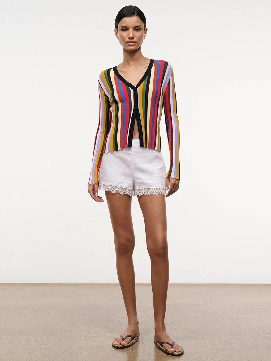 A woman stands indoors against a plain background wearing the STAUD Ezra Cardigan in Artist Stripe, paired with white lace-trimmed shorts and brown sandals.