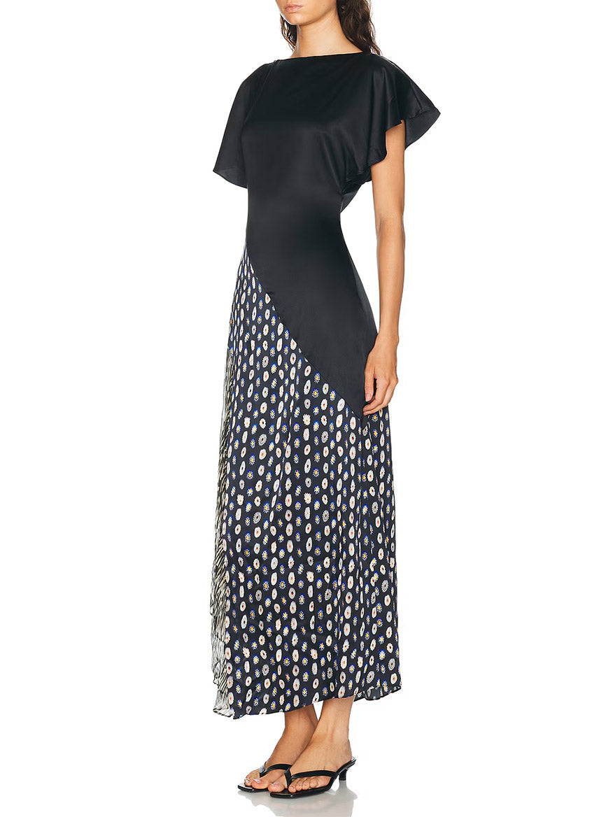 A woman wearing the STAUD Fawn Silk Dress in Black Multi—an elegant black short-sleeve handkerchief dress with an asymmetrical skirt and dark floral pattern, crafted from luxurious stretch silk—stands in sandals against a plain background.