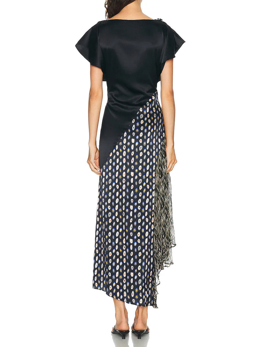 A woman stands facing away, wearing the STAUD Fawn Silk Dress in Black Multi—a black stretch silk dress with short sleeves, an asymmetrical skirt, and a pleated polka dot pattern.