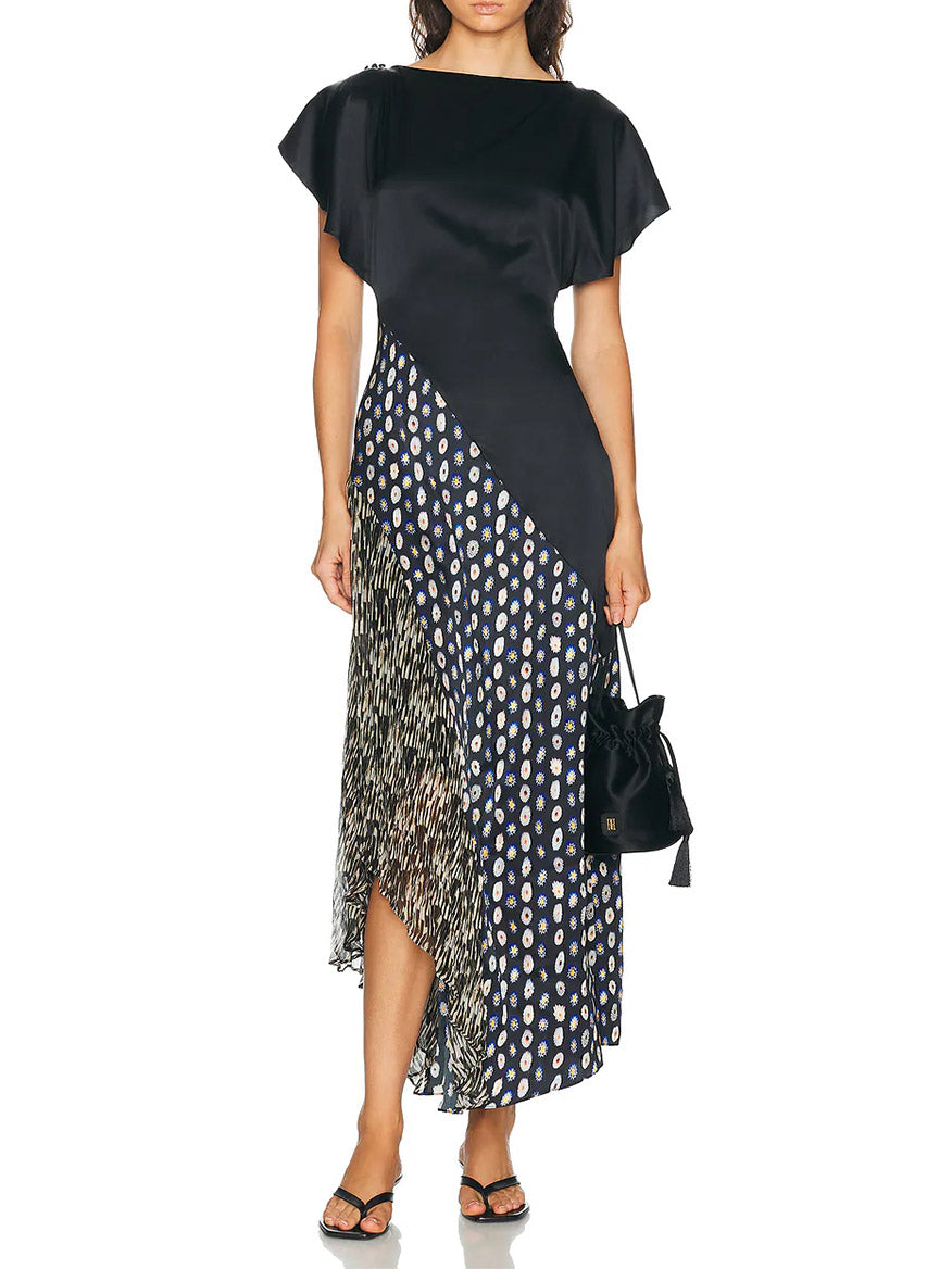 A woman wearing the STAUD Fawn Silk Dress in Black Multi—a short-sleeve, asymmetrical midi dress featuring polka dot and abstract patterns—paired with a black handbag and black sandals.