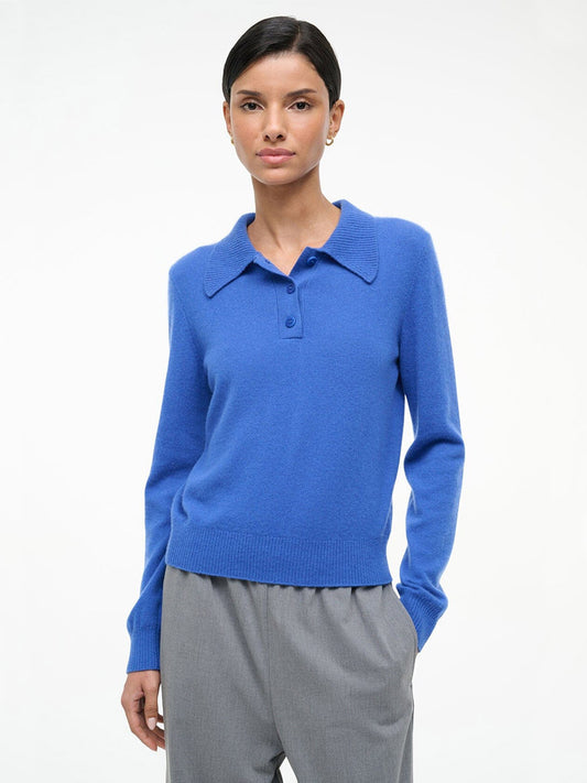 A person stands against a plain background wearing a STAUD Gemma Cashmere Sweater in Sapphire and gray trousers, with one hand in a pocket.