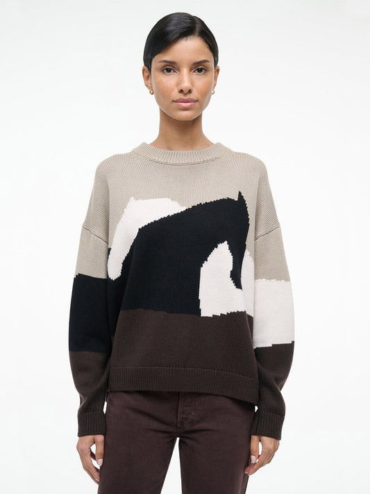 A person faces forward wearing the STAUD Giada Sweater in Stables, a relaxed-fit merino wool crewneck with abstract black, beige, white, and brown patterns, paired with dark brown pants against a plain white background.