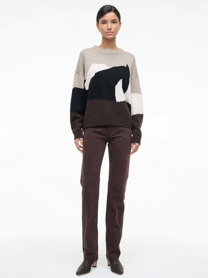 A woman stands against a plain background wearing the STAUD Giada Sweater in Stables, featuring a beige, black, and white abstract horse pattern, paired with dark brown pants and heeled shoes.