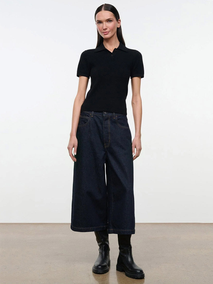 A woman wearing a black STAUD Gino Cashmere Polo Sweater with short sleeves, dark blue wide-leg jeans, and black boots.
