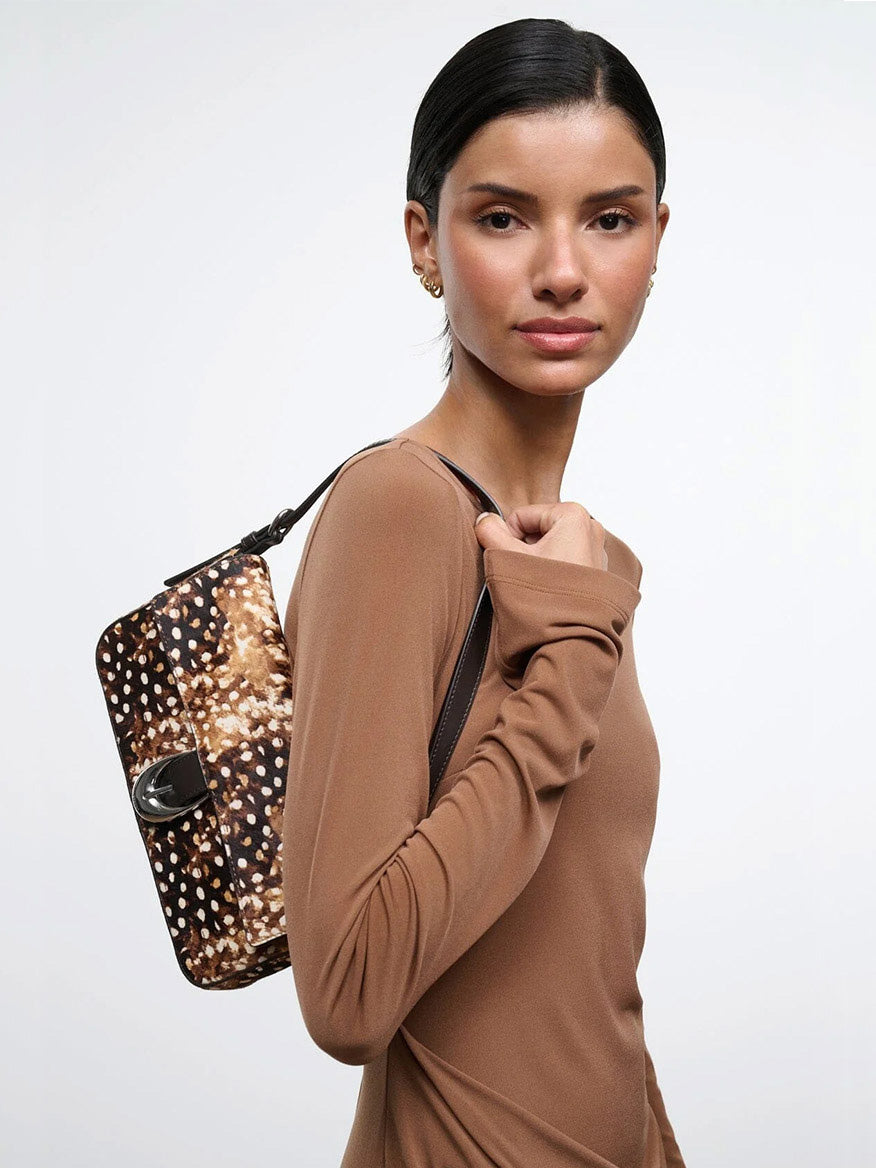 A woman in a tan long-sleeved top carries a STAUD Harlow Bag in Fawn Haircalf, which features brown and white spots and a silver buckle.