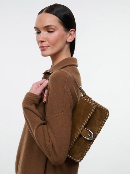 A person wearing a brown sweater carries a brown suede STAUD Harlow Bag in Cigar, featuring whipstitch detailing and a silver buckle closure.