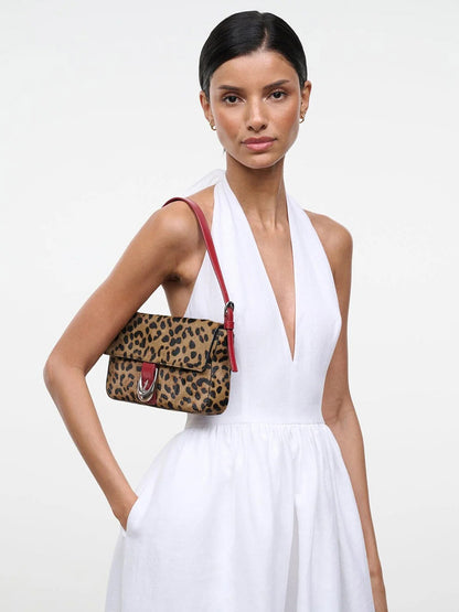 Woman in a sleeveless white dress stands with one hand in her pocket, carrying the STAUD Harlow Bag in Cheetah—a chic haircalf cheetah print handbag featuring a striking red strap and signature Moon buckle—over her shoulder against a plain background.