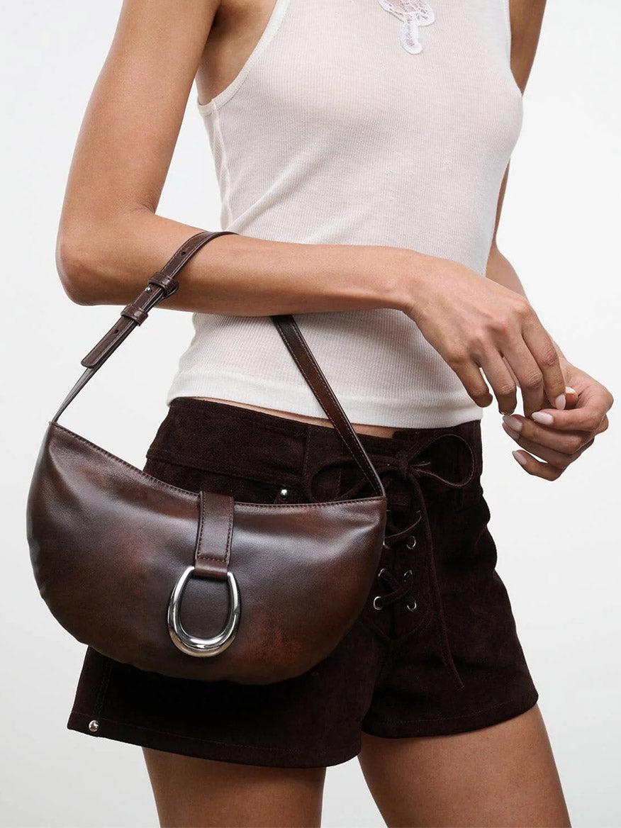 A person wearing a white tank top and brown shorts holds the STAUD Harlow Crescent Shoulder Bag in Espresso Distressed, featuring a striking silver moon buckle detail.
