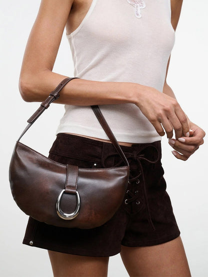 A person wearing a white tank top and brown shorts holds the STAUD Harlow Crescent Shoulder Bag in Espresso Distressed, featuring a striking silver moon buckle detail.