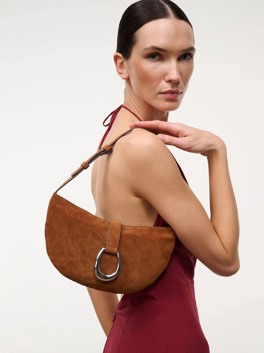 A person in a sleeveless maroon dress poses with the STAUD Harlow Crescent Shoulder Bag in Tabac, featuring a large moon buckle, against a plain light background.