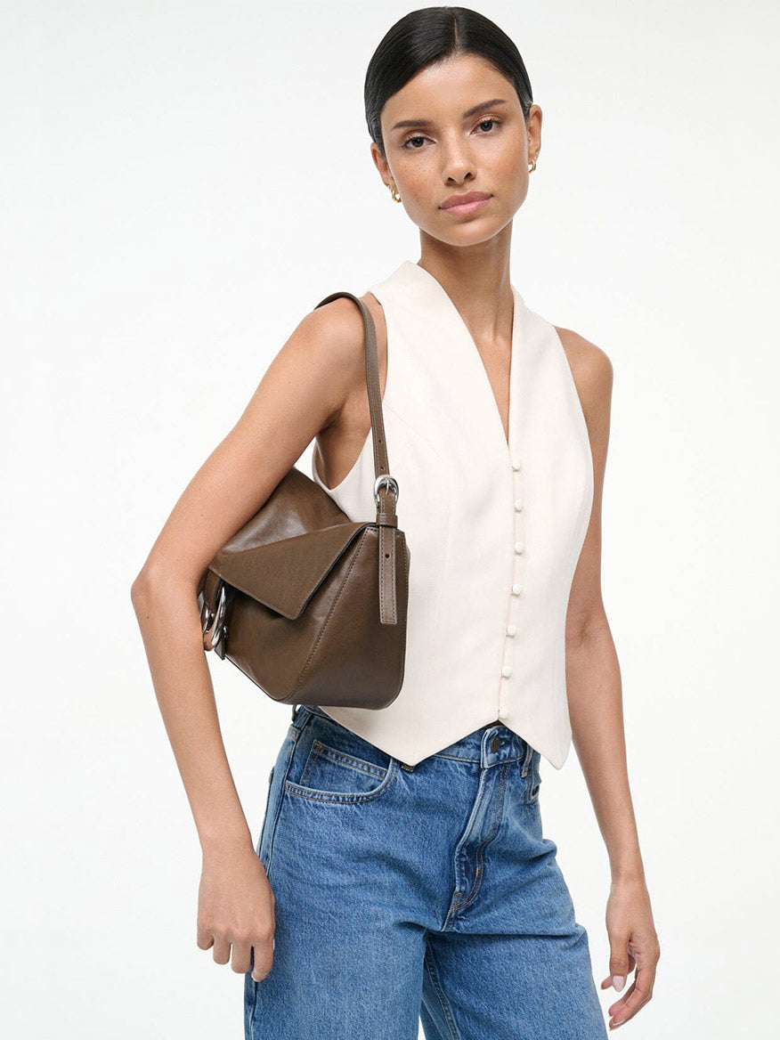 A person in a white sleeveless buttoned vest and blue jeans holds the STAUD Harlow Medium Bag in Truffle, a brown leather shoulder bag, against a plain white background.