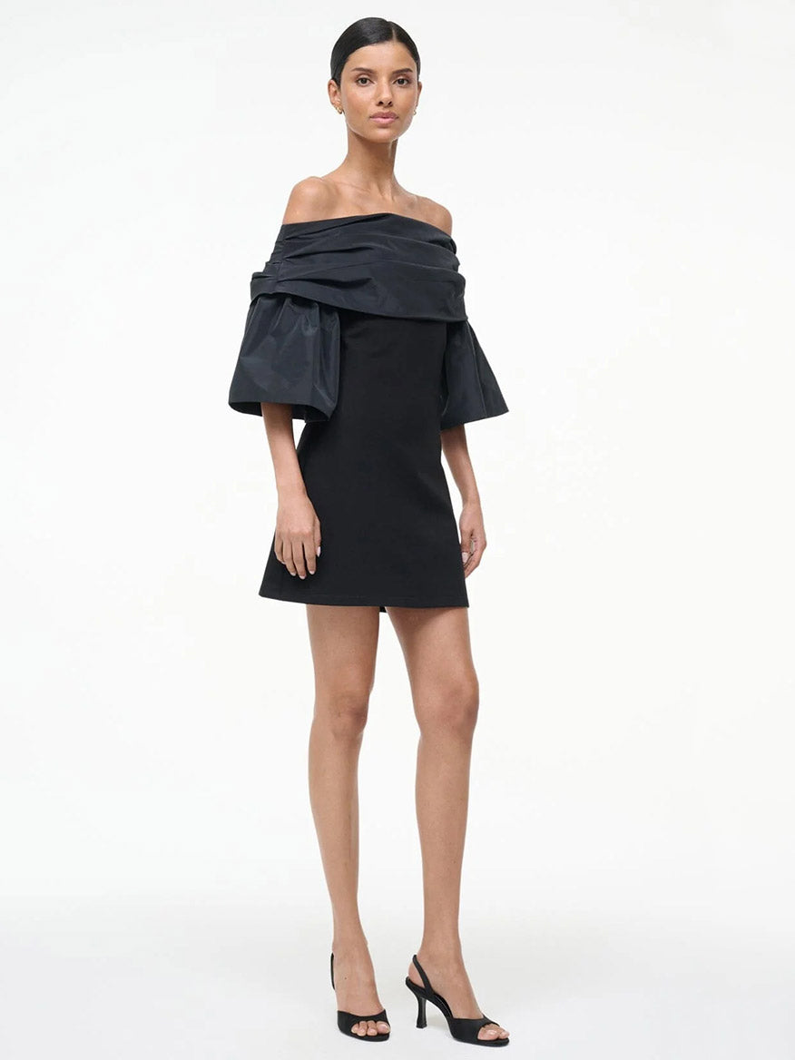 A woman poses against a white background wearing the STAUD Harper Dress in Black, featuring off-the-shoulder taffeta sleeves, paired with black open-toe heels.