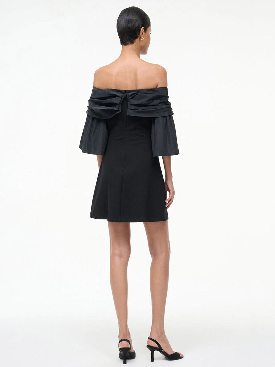 A person stands facing away in the STAUD Harper Dress in Black, featuring off-the-shoulder taffeta sleeves and paired with black heeled sandals, set against a plain white background.