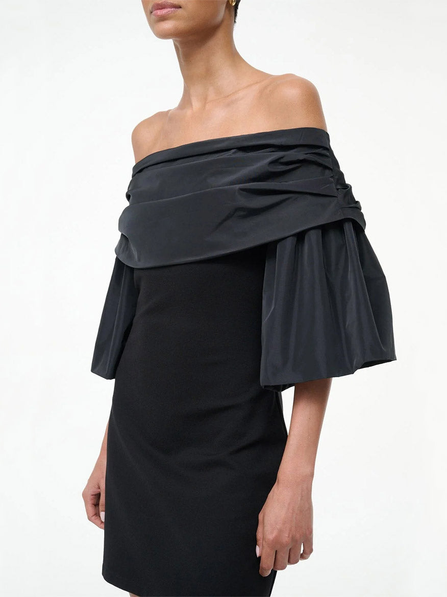 A woman poses against a plain white background wearing the STAUD Harper Dress in black, featuring off-the-shoulder styling and voluminous taffeta sleeves.