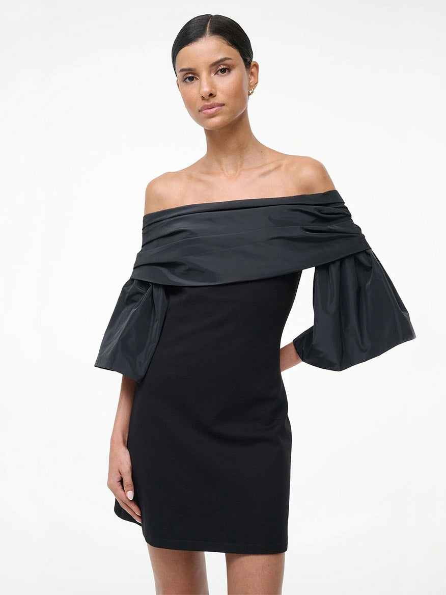 A woman wearing the STAUD Harper Dress in Black, featuring off-the-shoulder voluminous taffeta sleeves, stands against a plain white background.