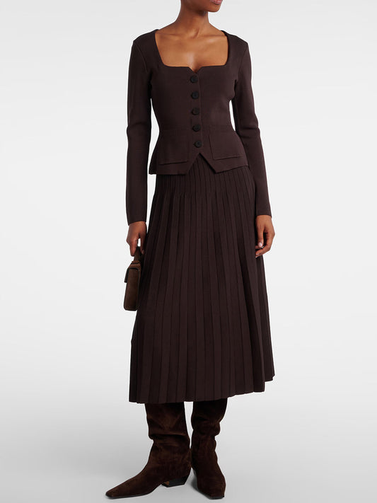 A person in a brown buttoned cardigan with a square neckline, the STAUD Lena Skirt in Earth featuring pleats and an elastic waistband, holding a small bag and wearing knee-high brown boots.
