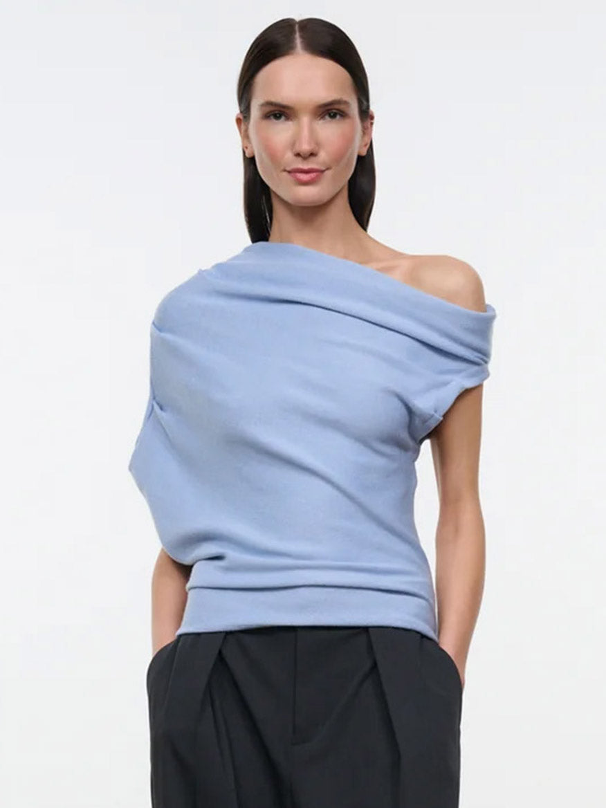 A model wears the STAUD Mabel Sweater in Powder Blue, a light blue, asymmetrical knit top featuring draped detailing on one shoulder.