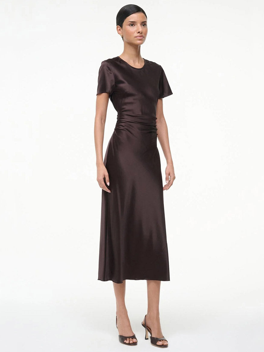 A woman stands against a plain white background wearing the STAUD Marcel Silk Dress in Earth, a short-sleeve, dark brown midi dress, paired with black open-toe heels.