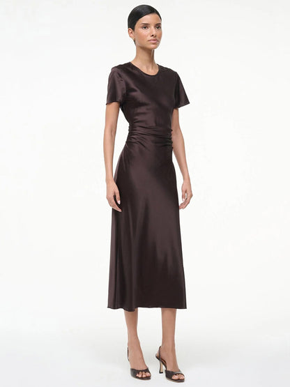 A woman stands against a plain white background wearing the STAUD Marcel Silk Dress in Earth, a short-sleeve, dark brown midi dress, paired with black open-toe heels.
