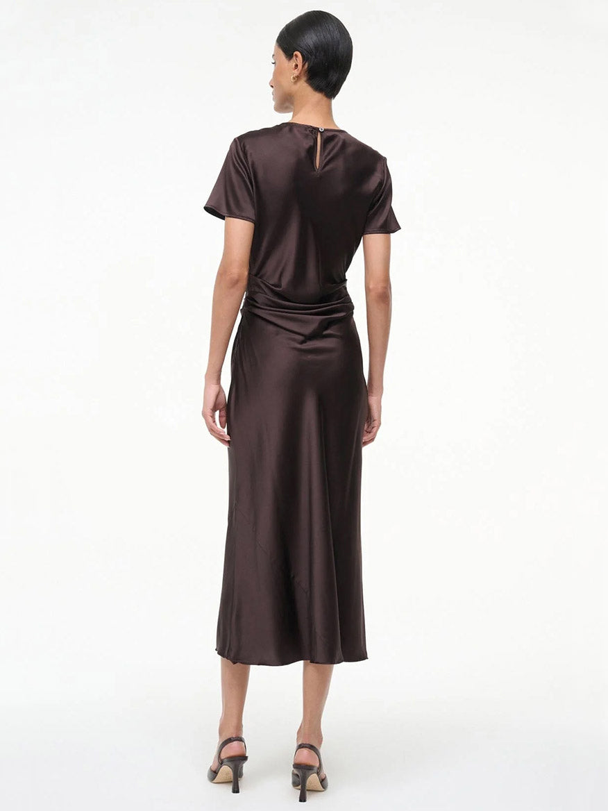 A woman stands facing away, wearing the STAUD Marcel Silk Dress in Earth with short sleeves and open-toe heeled sandals.
