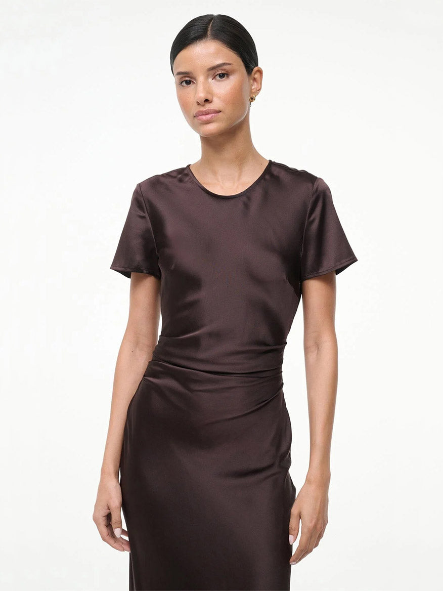 A woman with dark hair in a low bun wears the STAUD Marcel Silk Dress in Earth, a short-sleeve, dark brown cocktail dress, and stands against a plain white background.
