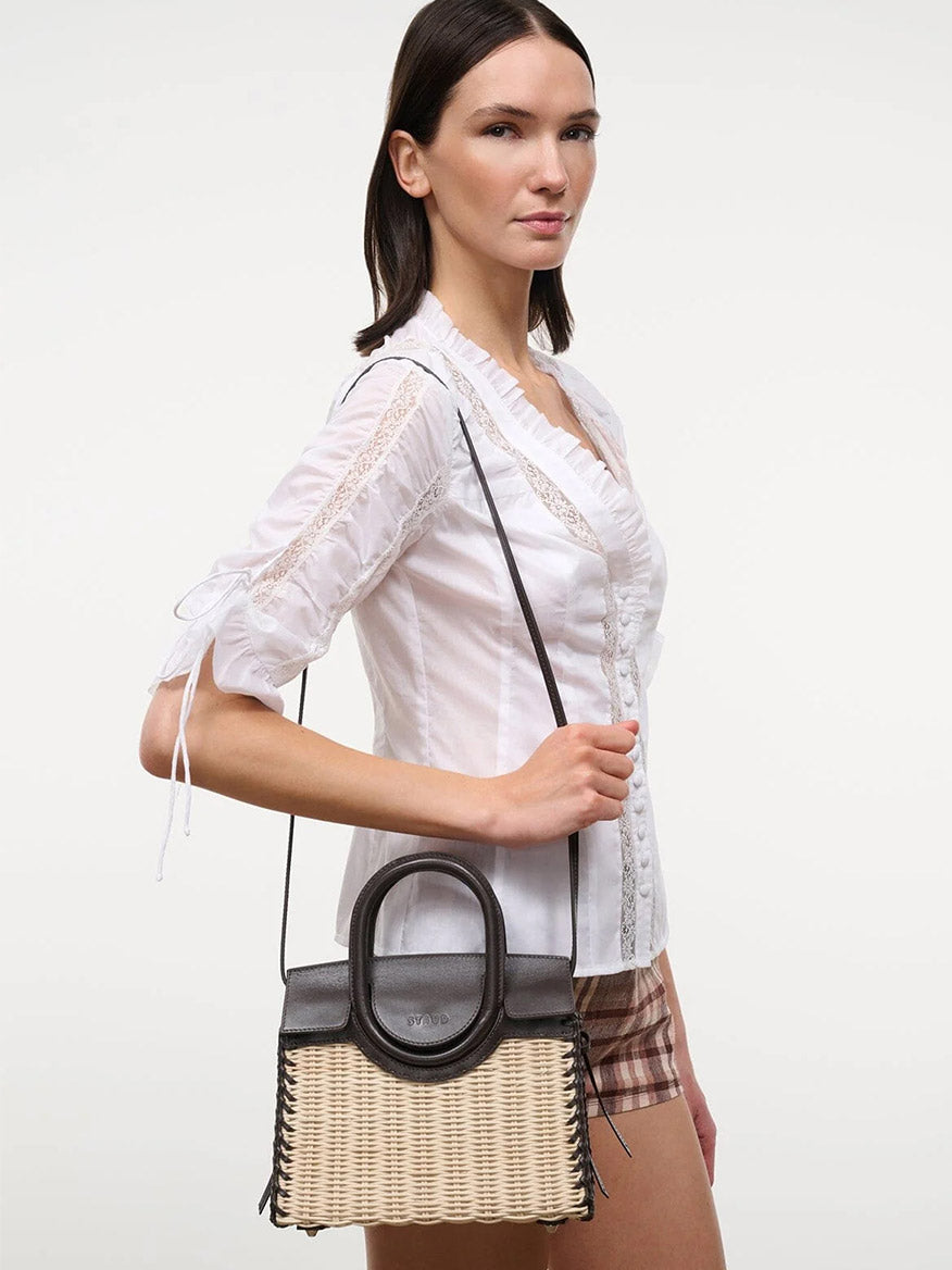 A woman in a white blouse and plaid shorts holds the STAUD Maude Mini Wicker Carryall in Natural/Espresso with black leather accents against a plain background.
