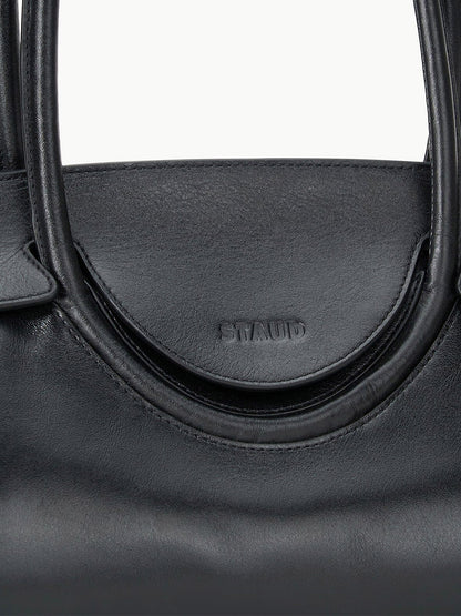 Close-up of the STAUD Maude Shoulder Bag in Black, crafted from polished leather with rounded handles, an east-west silhouette, and "STAUD" embossed on the front flap.