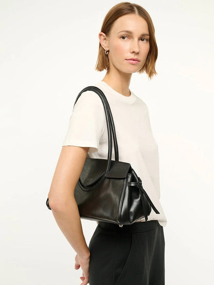 A woman with straight, shoulder-length hair wears a white t-shirt and black pants, holding the STAUD Maude Shoulder Bag in Black against a plain background.