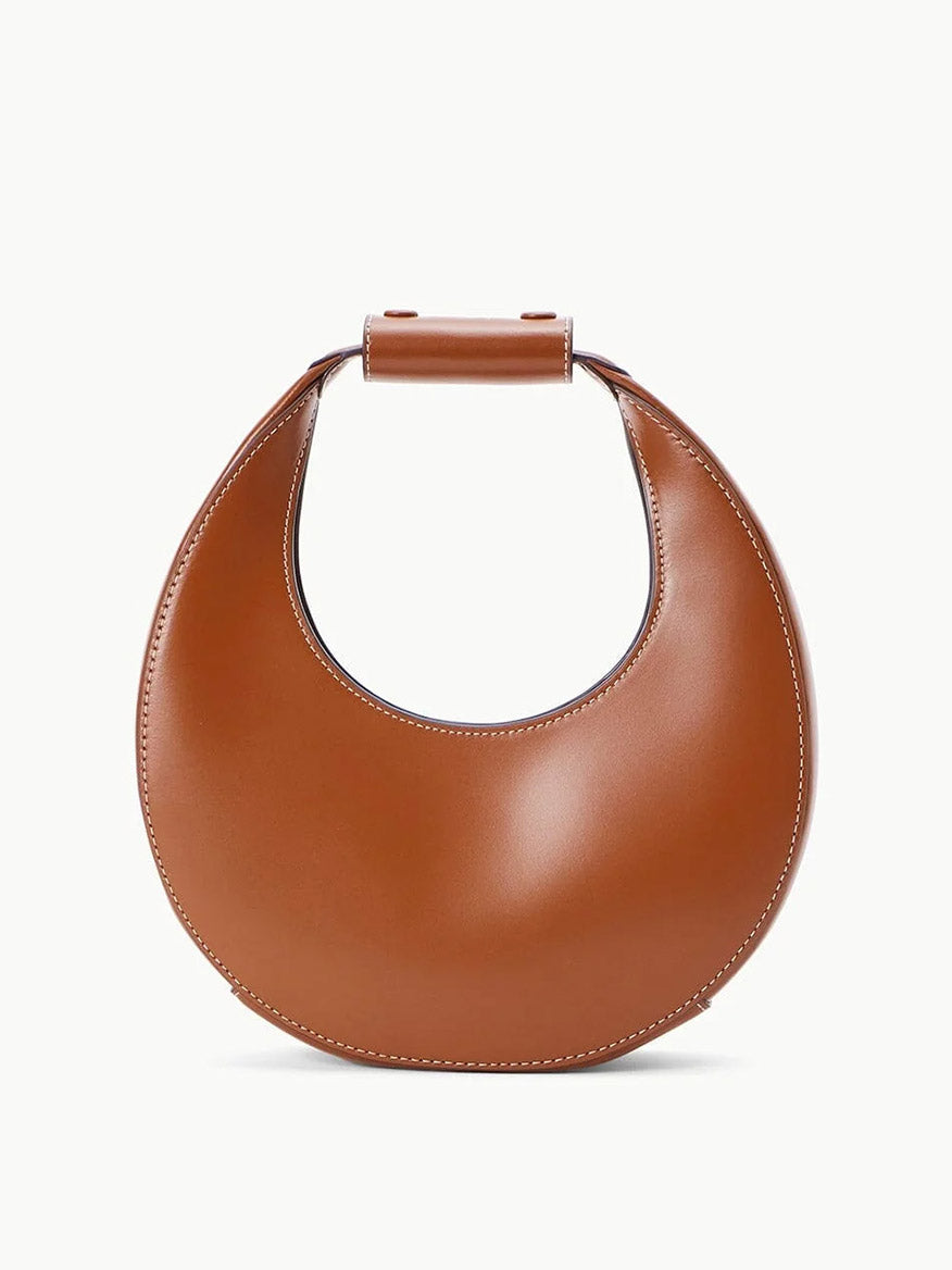 The STAUD Mini Moon Tote Bag in Tan boasts a distinctive crescent moon shape, crafted in brown leather with noticeable stitching and a rounded handle. Its versatility shines through with the addition of an optional crossbody strap, complementing its light background.