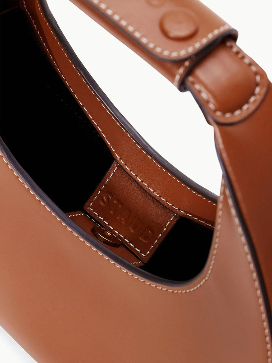 Close-up of a STAUD Mini Moon Tote Bag in Tan, featuring white stitching, a crossbody strap, and highlighting part of the crescent-shaped interior with a visible brand patch.