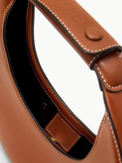 Close-up of the STAUD Moon Tote Bag in Tan, crafted in crescent moon shape with brown leather. It features a strap with a button closure, visible stitching, and is complete with luxurious suede lining.